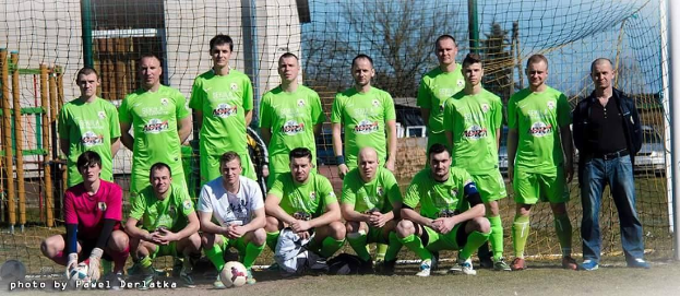 Mannergruppe in grünen T-Shirts auf einem Fußballfeld mit Ball und Torpfosten, Bäume und Gebäude im Hintergrund.