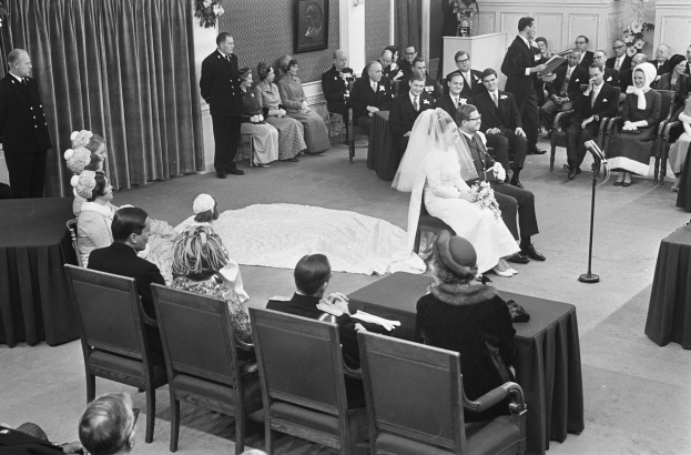 Schwarzes und weißes Foto einer Hochzeitszeremonie mit sitzenden und stehenden Menschen, einem Mann, der in ein Mikrofon spricht, und einer Frau mit einem Blumenstrauß, mit einem Podium, Blumengestecken, einem Vorhang, einem gerahmten Foto an der Wand und einer Tür im Hintergrund.