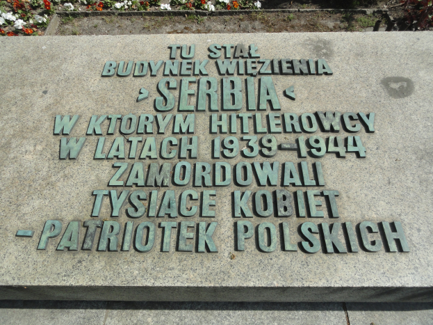 Gedenkstein mit Inschrifttafel vor einem Gebäude, umgeben von blühenden Pflanzen.