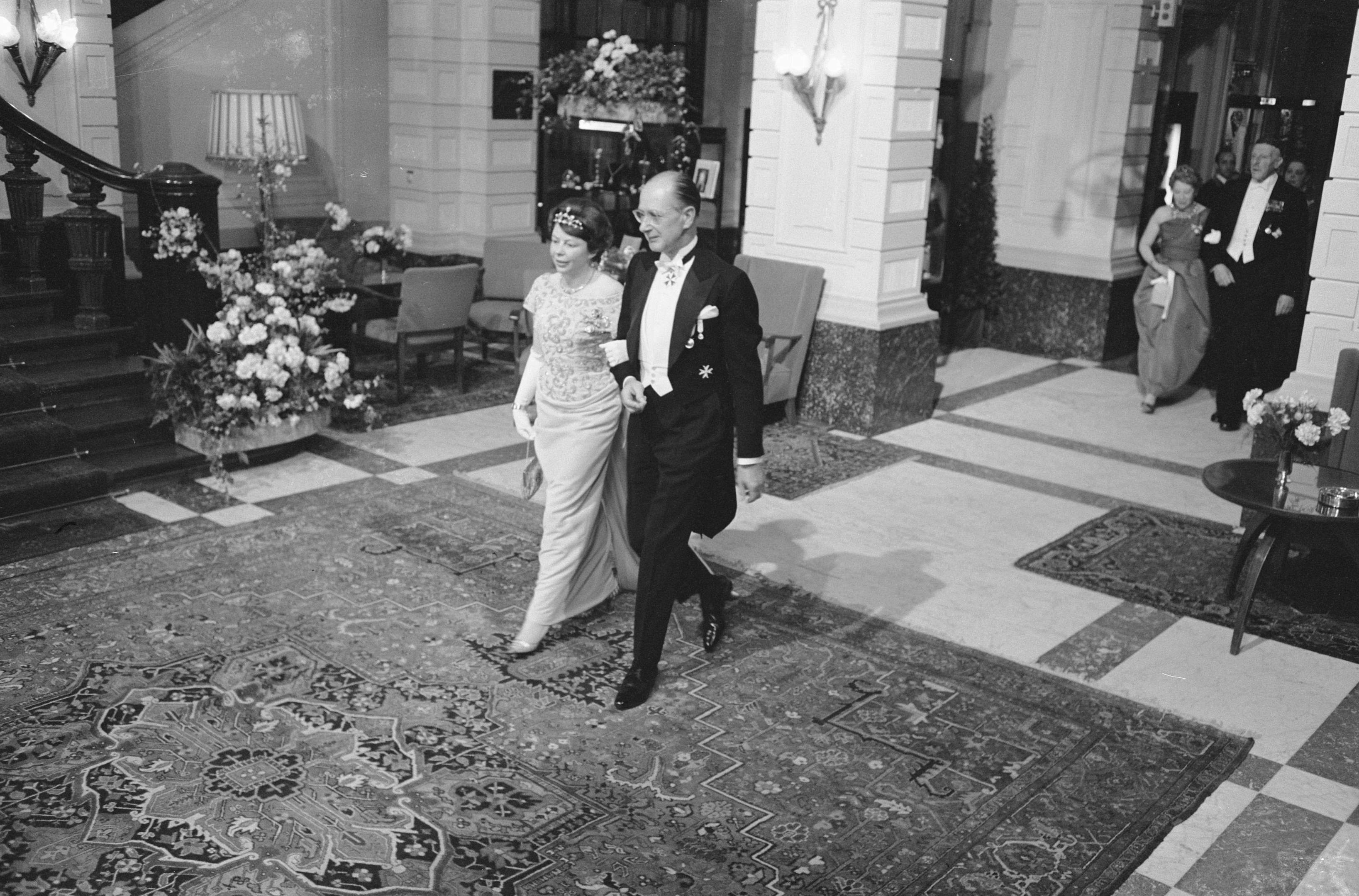 Schwarzes und weißes Foto einer Braut und eines Bräutigams, die durch die Hotellobby gehen, umgeben von Gästen, einem Blumenstrauß auf einem Tisch, Stühlen, Topfpflanzen mit Blüten, Lampen, einer Treppe mit Geländer und einer Wand.