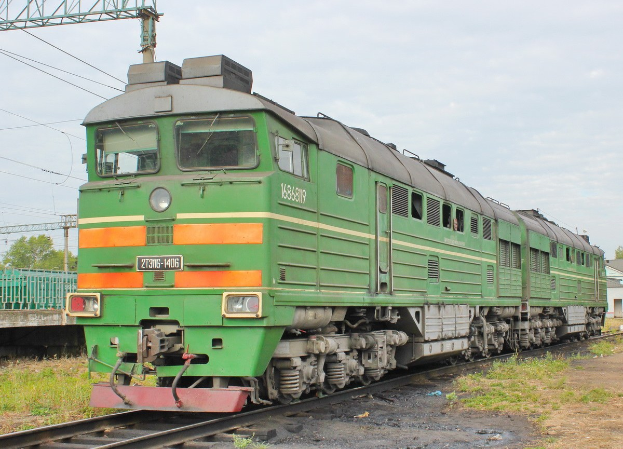 Ein grüner Diesel-Elektrolokomotive fährt auf Eisenbahnschienen durch einen bewaldeten Bereich, mit einer Brücke und einem Haus im Hintergrund, unter einem bewölkten Himmel.