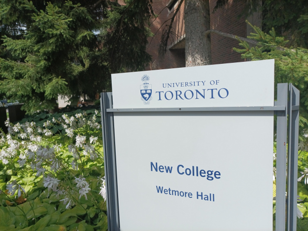 Außenansicht des neuen Colleges der Universität Toronto an Wetmore Hall, mit einem Schild vor dem Gebäude, umgeben von grünem Gras, bunten Blumen, hohen Bäumen und einem hellblauen Himmel.
