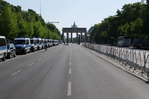 Eine Reihe von Polizeiwagen, die auf der Seite einer Straße vor dem Brandenburger Tor in Berlin, Deutschland, geparkt sind, mit Menschen, die Fahrräder fahren und auf der Straße stehen, Barrieren und Bäume, die die Seiten säumen, und einem Bogen mit Statuen im Hintergrund.
