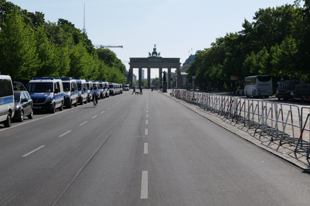 Eine Reihe von Polizeiwagen, die auf der Seite einer Straße vor dem Brandenburger Tor in Berlin, Deutschland, geparkt sind, mit Menschen, die Fahrräder fahren und auf der Straße stehen, Barrieren und Bäume, die die Seiten säumen, und einem Bogen mit Statuen im Hintergrund.
