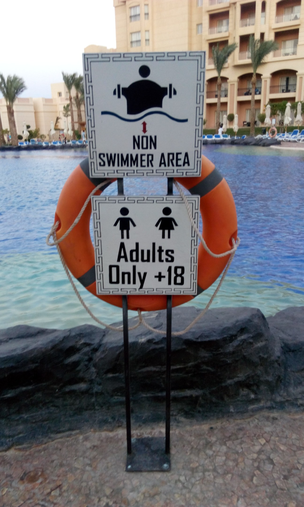 Ein Schwimmbad mit einem "Nichtschwimmerbereich Erwachsene nur"-Schild im Vordergrund, umgeben von Bäumen, Gebäuden und einem klaren blauen Himmel, mit einem Rettungsring in der Mitte des Pools.