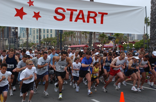 Gruppe von Läufern bei einem Marathon, die an einer Verkehrskegel vorbeilaufen, mit einem Banner und Gebäuden im Hintergrund bei klarem Himmel.