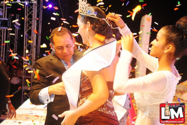 Ein Mädchen auf der Bühne bei einer Miss-World-Show mit einer Krone, mit einem anderen Mädchen, das sie hinter ihr anpasst, und einem Mann in einem schwarzen Anzug, der vor ihr die Hand schüttelt, vor einem Hintergrund aus Metallrahmen und einer dunklen Hintergrund.