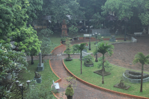 Ein grüner Park mit Bäumen, Pflanzen, Gras, Laternenmasten, Bänken und Skulpturen, mit einem Gebäude im Hintergrund.