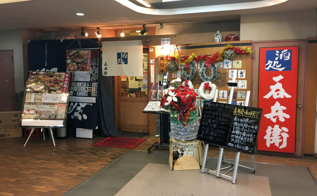 Eingangsbereich eines japanischen Restaurants mit Regalen, einer Vase, einer Texttafel auf einem Ständer, einem Teppich, einer Pappkiste, Papieren an der Wand, Deckenleuchten, einem Vorhang, einer Tür und einem Schild auf der rechten Seite.