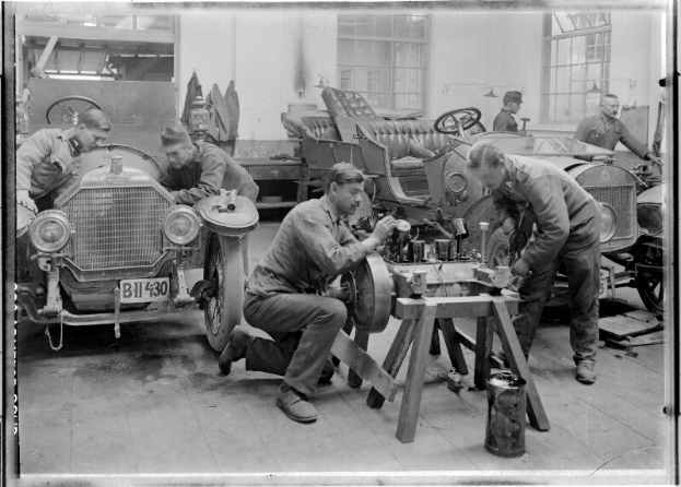 Eine Gruppe uniformierter Männer, die an einem Auto in einer schwarzen und weißen Garage arbeiten, umgeben von Werkzeugen und Equipment auf einem zentralen Schreibtisch.