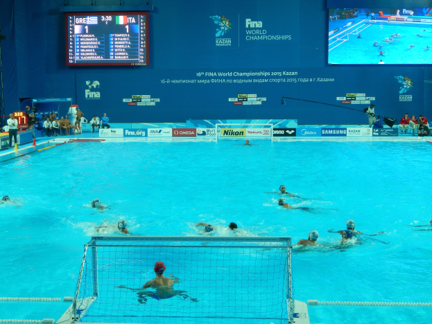 Eine Gruppe von Menschen spielt Wasserball in einem großen Pool mit einem Netz, Zuschauern auf der linken Seite und einer Wand mit Text und Logos im Hintergrund bei den FINA-Weltmeisterschaften 2016 in Kazan.