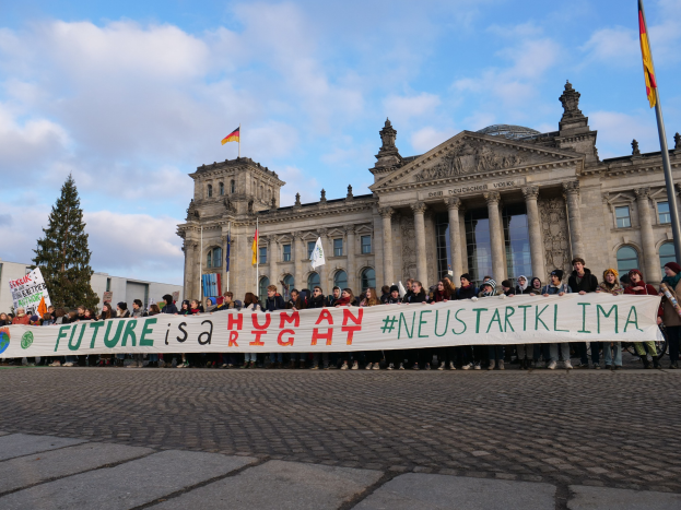 Gruppe von Menschen mit einem Transparent mit der Aufschrift 'Zukunft ist ein Menschenrecht' vor dem Reichstag in Berlin, mit Bäumen und Fahnenmasten im Hintergrund.