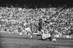 Schwarze und weiße Fotografie von Männern, die auf einem Feld Fußball spielen, mit Zuschauern im Hintergrund und einem Banner "Bundesliga 1978-79 Ajax 1-1 Hannover 96".
