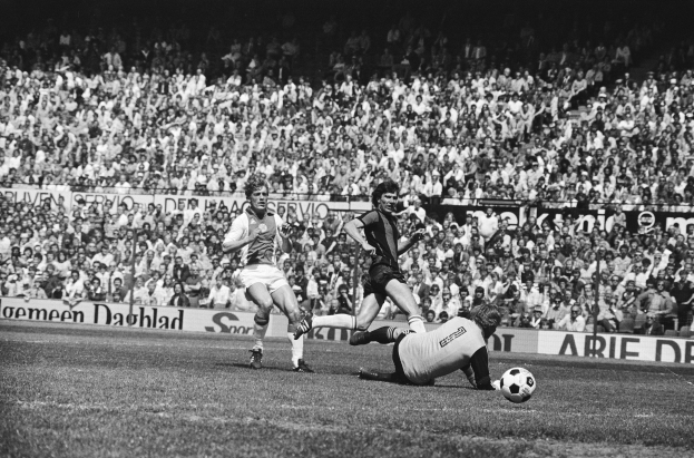 Schwarzes Foto von Männern, die auf einem Feld Füßball spielen, mit Zuschauern im Hintergrund und einem Banner mit der Aufschrift "Bundesliga 1978-79 Ajax 1-1 Hannover 96."