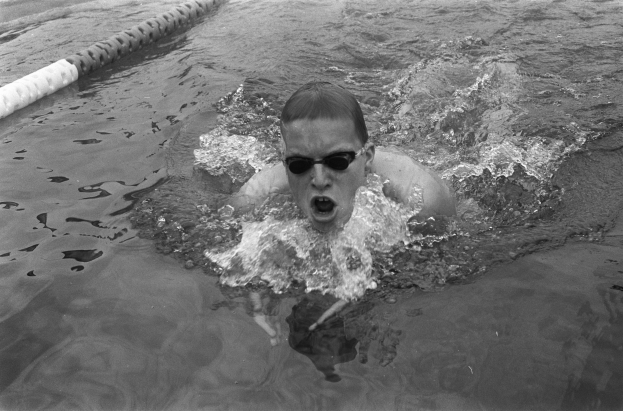 Ein Mann schwimmt in einem Pool, trägt eine Schwimmbrille und hat den Mund offen, umgeben von Bahnmarkierungen, auf einem Schwarz-Weiß-Bild.