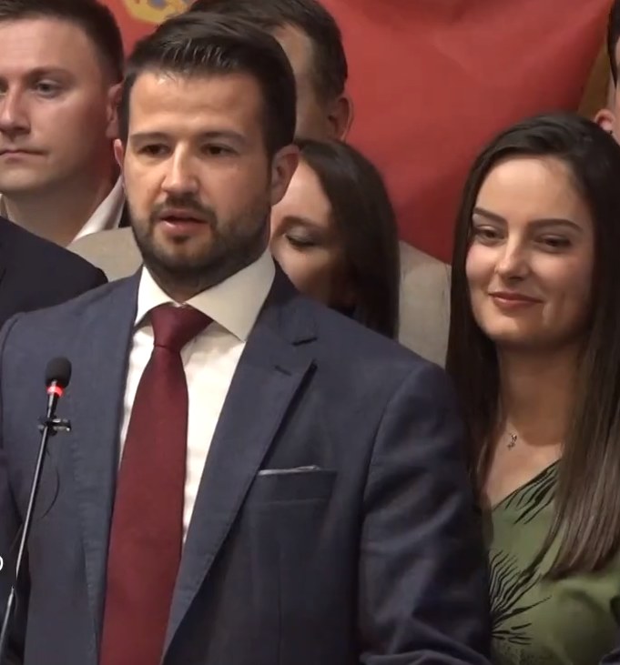 Ein Mann im Anzug spricht in ein Mikrofon an einem Rednerpult mit einer Menge davor und einem Banner dahinter, wahrscheinlich zu einem Regierungssozialmedienverbot.