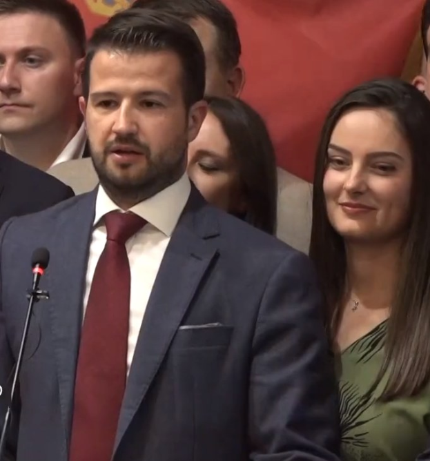 Ein Mann im Anzug spricht in ein Mikrofon an einem Rednerpult mit einer Menge davor und einem Banner dahinter, wahrscheinlich zu einem Regierungssozialmedienverbot.