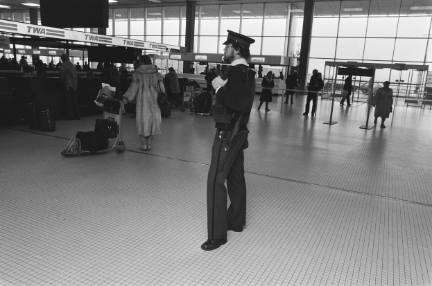 Polizeibeamter in Uniform mit Waffe in einem Flughafen-Terminal umgeben von Menschen und Gepäck mit Trolleys, Deckenleuchten, Pfosten und Glaswänden im Hintergrund.