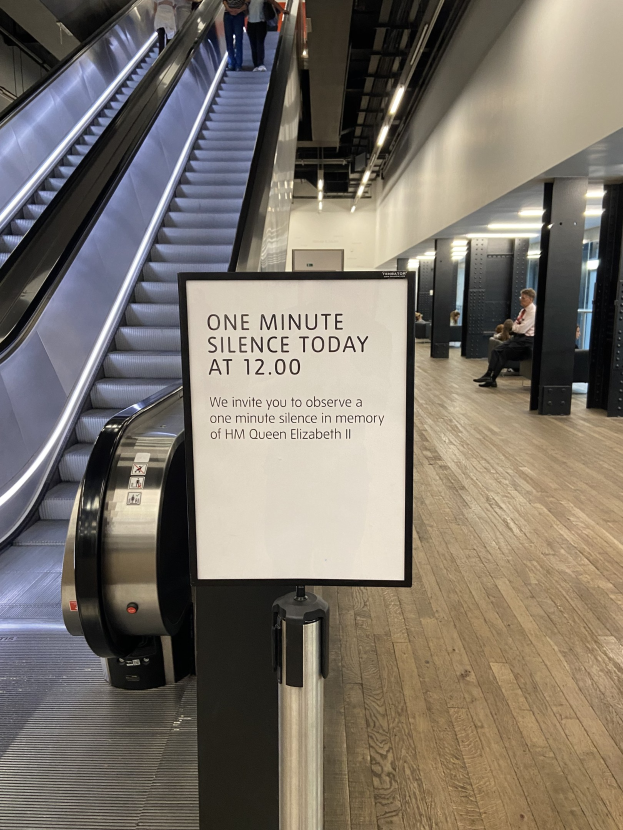 Eine Rolltreppe im Flughafen mit einem Schild, auf dem "Eine Minute Stille heute" steht, einige Menschen darauf und an der Decke befestigte Lampen im Hintergrund.