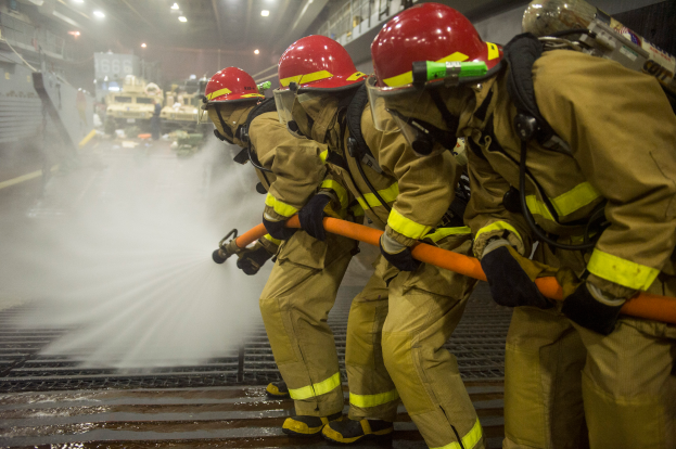 Feuerwehrleute in Helmen und Handschuhen spritzen Wasser aus Rohren auf ein Feuerwehrauto, mit verschiedenen Gegenständen und einem Boden im Hintergrund.