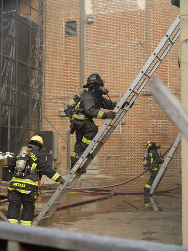 Feuerwehrleute in Helmen und Ausrüstung klettern an einer Leiter vor einem Backsteingebäude mit Rohren auf dem Boden und einem Metallstab unten, mit einem anderen Gebäude und einem Netz im Hintergrund.