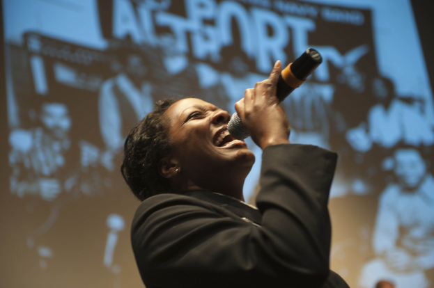 Eine Frau in einem schwarzen Kleid mit einem Dutt singt leidenschaftlich in ein Mikrofon auf der Bühne, ihr Gesicht von Scheinwerfern beleuchtet, die Augen vor Emotion geschlossen.