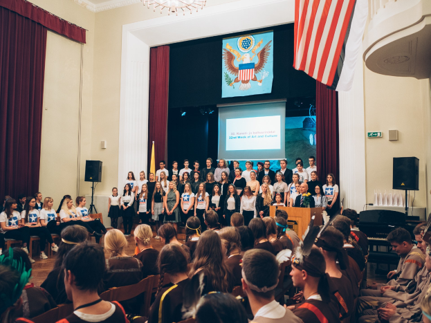 Eine Gruppe von Menschen steht auf einem Podium mit Mikrofon, flankiert von Fahnen und Lautsprechern, vor einem sitzenden Publikum.