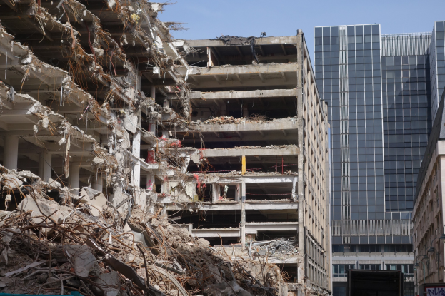 Große Abrissarbeiten in einer Stadt, umgeben von intakten Gebäuden, Straßeninfrastruktur, Fahrzeugen, Bäumen und einem klaren Himmel.