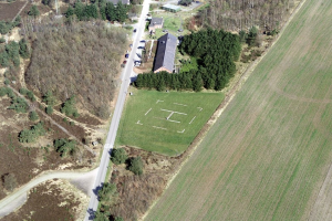 Eine Luftaufnahme eines Bauernhofs in einem Feld, umgeben von Bäumen, Häusern, Fahrzeugen, Straßen und Gras.