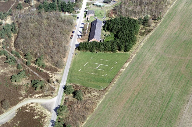 Eine Luftaufnahme eines Bauernhofs in einem Feld, umgeben von Bäumen, Häusern, Fahrzeugen, Straßen und Gras.