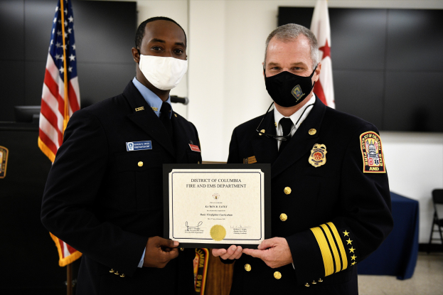 Zwei Männer in Feuerwehrausstattung stehen nebeneinander und tragen Masken sowie ein Zertifikat; im Hintergrund sind Flaggen und Möbel zu sehen; der Mann links ist der Feuerwehrchef des Bezirks Washington und erhält eine Auszeichnung.