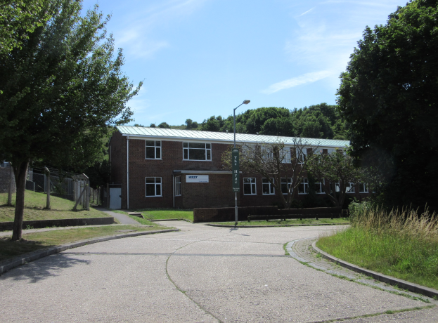 Ein Schulgebäude mit Fenstern, ein Straßenmast, ein Schild, eine Baumgruppe, Gras, ein Zaun und ein bewölkter Himmel, das sich in der Mitte eines Parks mit saftigem Gras befindet.