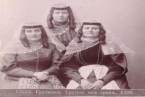 Drei Frauen in traditionellen russischen Hochzeitskleidern mit aufwändiger Stickerei und Perlen stehen vor einer weißen Wand in einem Schwarz-Weiß-Foto.