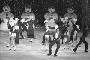 Eine Gruppe von Menschen in Kostümen fährt auf einer Eisbahn im Schnee Schlittschuh, mit Schneemännern im Hintergrund, in Schwarz-Weiß aufgenommen.