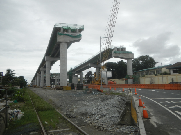 Baustelle mit einer Brücke im Hintergrund, Straße mit Verkehrskegeln markiert, Bahnschiene links, verstreute Steine und Gras, Bäume und Gebäude säumen die Straße und ein bewölkter Himmel.