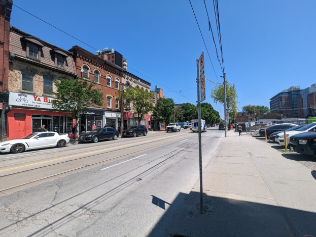 Stadtstraße mit parkenden Autos, Gebäuden, Bäumen, Strommasten und einem Schild "New York City Immobilien zum Verkauf" vor einem Himmelhintergrund.