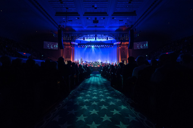 Ein großer Auditorium mit sitzenden Zuschauern, eine Bühne mit Musikern und Deckenbeleuchtung und Fernseher, was auf ein Konzerthaus während der Nationalkonvention 2019 hinweist.