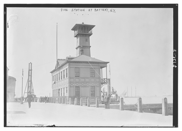 Schwarz-weiß-Foto einer Feuerwache an der Battery, New York, mit einem Gebäude, Menschen, Pfählen, einem Zaun, Booten auf dem Wasser und einem bewölkten Himmel.