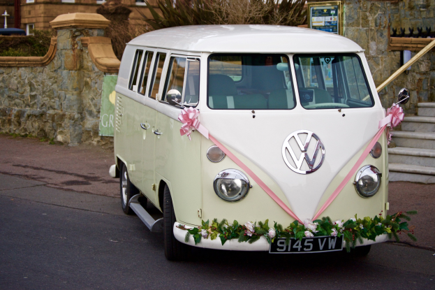 Ein weißer und pinkfarbener VW-Campervan mit floralen Verzierungen, der vor einer Steinmauer geparkt ist, mit einer Treppe auf der rechten Seite und einem Gebäude im Hintergrund.