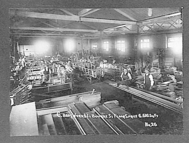 Schwarzes und weißes Foto von Arbeitern in einer Fabrik, die hölzerne Gegenstände zusammenbauen, mit sichtbaren Lichtern und Fenstern und einem Text unten, der "C. Body Works, St. Bourne's Floor Space, No. 26" lautet.