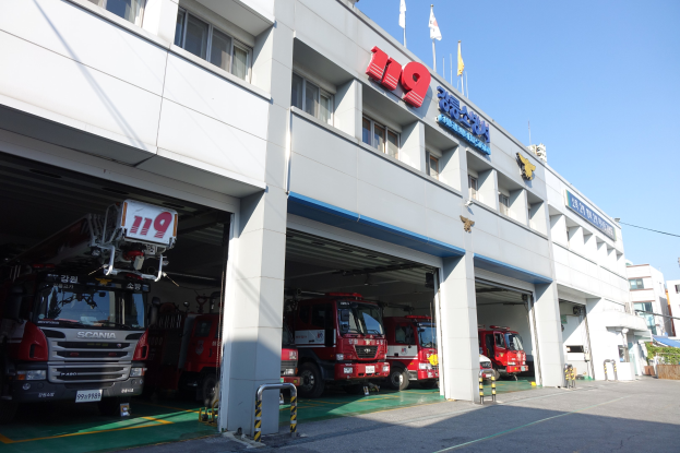 Feuerwache mit mehreren Feuerwehrautos vor der Tür, mit Fenstern, Säulen und Namensschildern, sowie Fahnen im Vordergrund und anderen Gebäuden gegen einen klaren blauen Himmel.