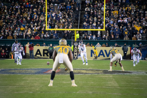 Ein Fußballspiel in vollem Gange mit Spielern in Helmen und Sportbekleidung im Vordergrund und einer Zuschauermenge im Hintergrund, mit sichtbaren Pfählen und Tafeln mit Text.