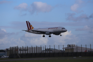 Germanwings Airbus A320-200 startet am Frankfurter Flughafen, mit Gras, einem Zaun, Gebäuden, Bäumen und einem bewölkten Himmel im Hintergrund.