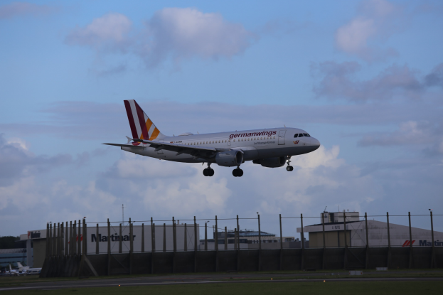 Germanwings Airbus A320-200 startet am Frankfurter Flughafen, mit Gras, einem Zaun, Gebäuden, Bäumen und einem bewölkten Himmel im Hintergrund.