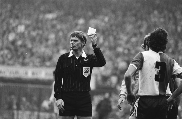 Schwarzes und weißes Bild eines Schiedsrichters, der eine rote Karte in einer Menge von Fußballspielern hält, mit einem Zaun im Hintergrund.