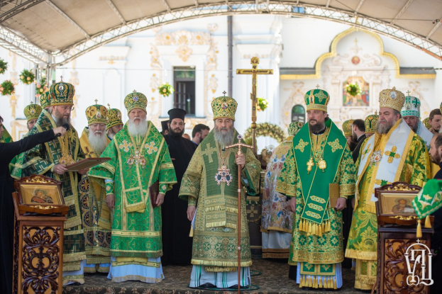 Eine Gruppe von Priestern vor einer Kirche, mit einem Buch und einem Mikrofon haltend, während einer religiösen Zeremonie mit einem Kreuz, Blumen und einem Gebäude im Hintergrund.