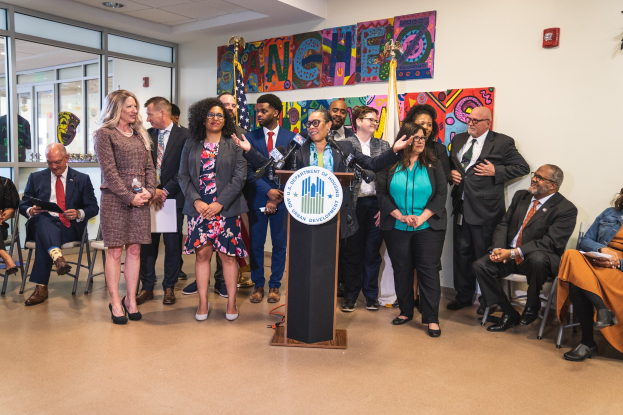 Gruppe von Menschen in einem Raum mit einem Podium, einem Mikrofon und einer Tafel, die "Sancho City Councilman Joins City Council" anzeigt, umgeben von Stühlen, Flaggen und Deckenleuchten.