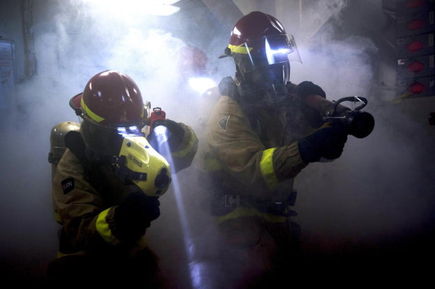 Zwei Feuerwehrleute in Schutzausrüstung sprühen Wasser auf eine rauchgefüllte Wand, mit einer Tafel und verstreuten Gegenständen in der Nähe.