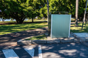 Transportable Toilette am Straßenrand in einem Park, umgeben von Bäumen, Gras, einem Gewässer und einem klaren blauen Himmel.