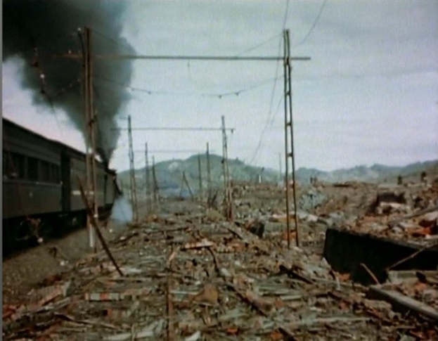 Ein Zug fährt durch eine verwöstete Landschaft mit aufsteigendem Rauch, umgeben von Strommästen und Dröhten, vor Höhen unter einem klaren Himmel, mit verstreuten Trömmern im Vordergrund.